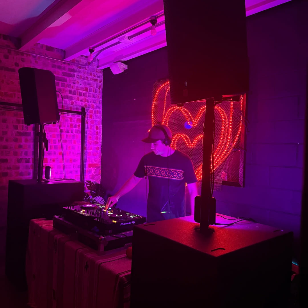 A DJ performs in front of a neon heart sign under purple lighting