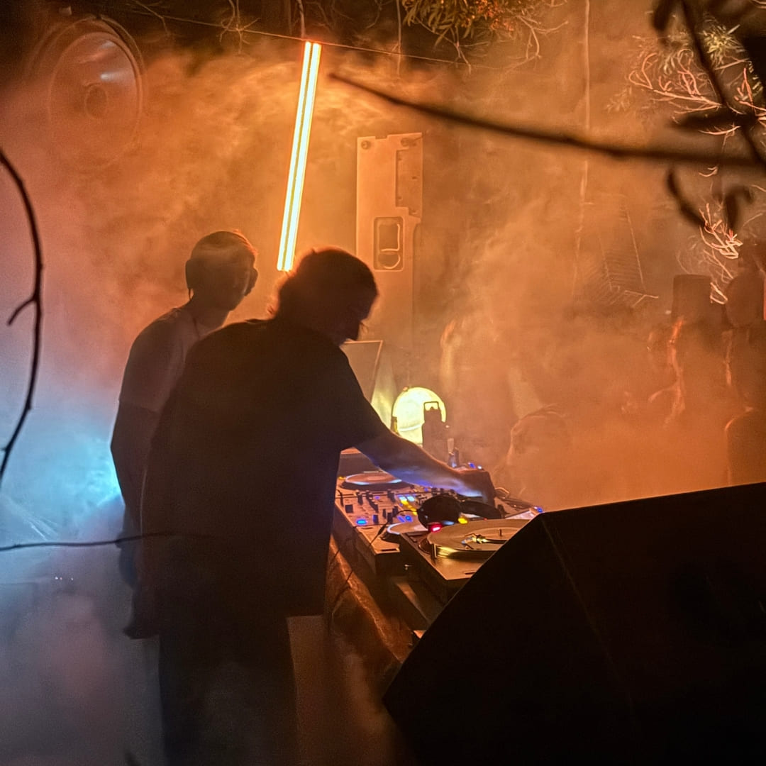 A DJ performs behind decks in silhouette with a glowing light bar and smoke effects