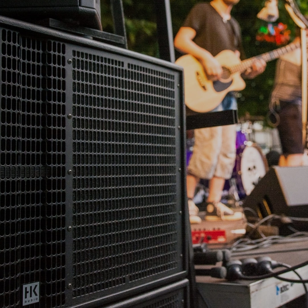 A professional HK Audio speaker in the foreground with a guitarist performing behind it