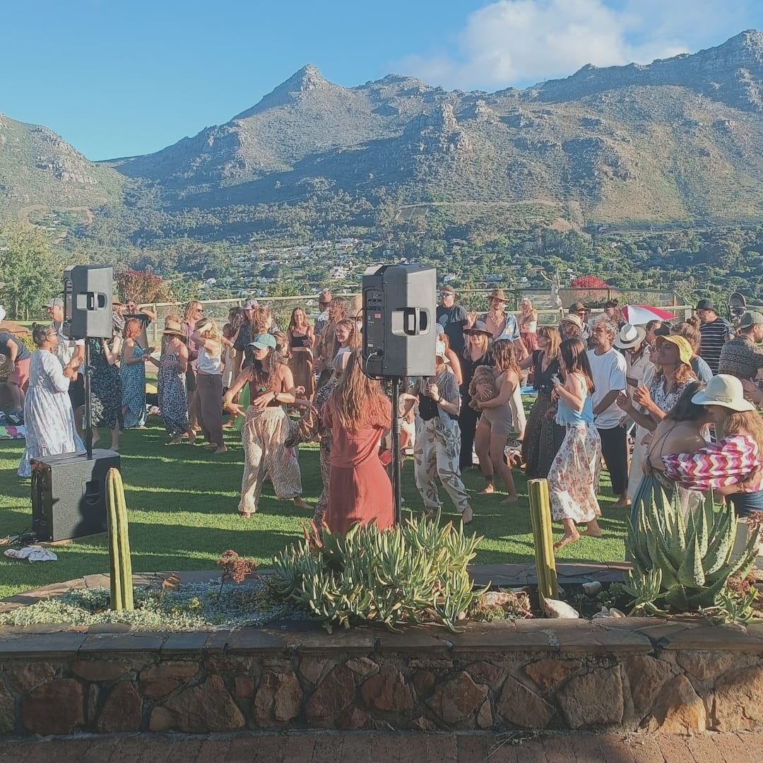 A crowd dances outdoors with mountains in the background and speakers set up on stands