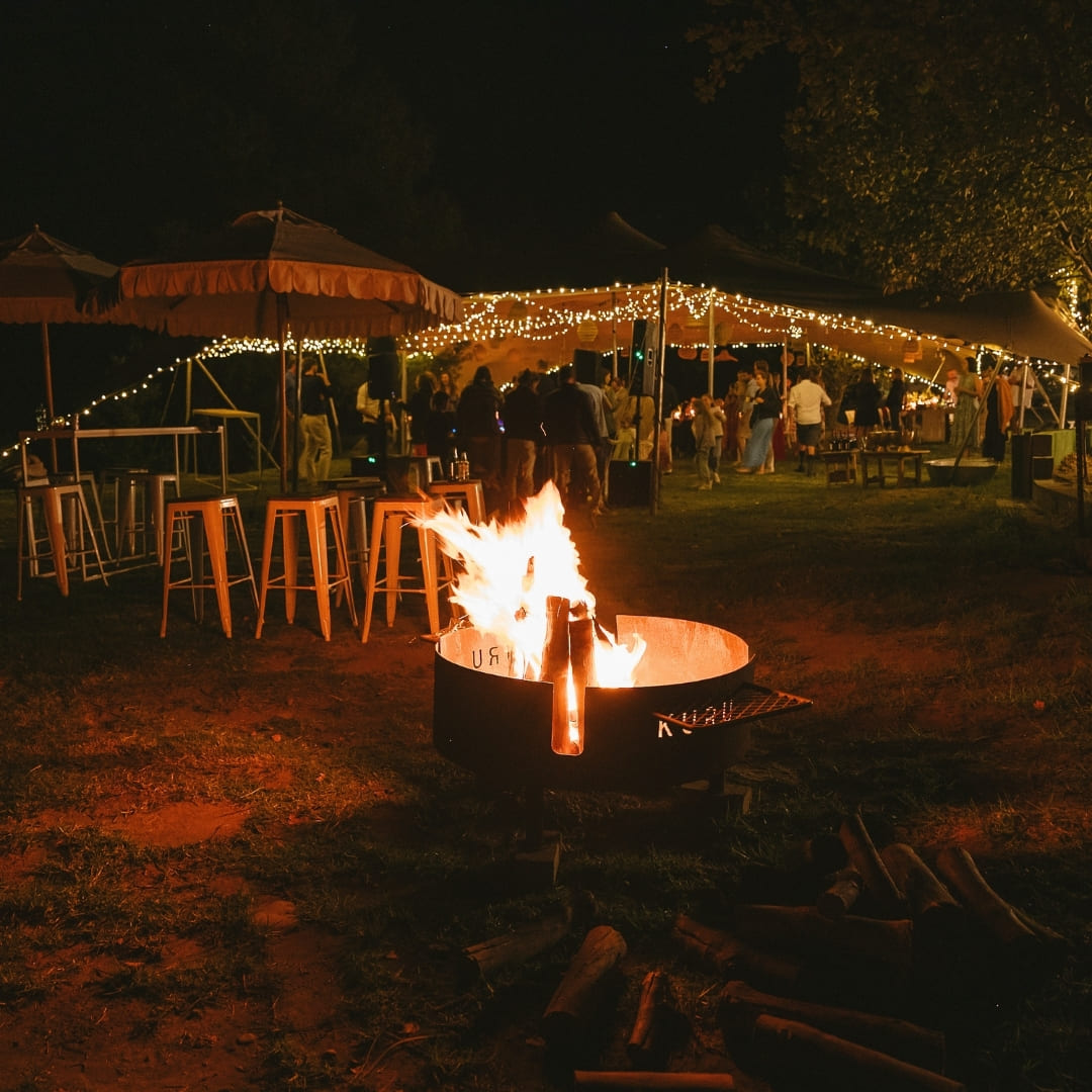 A canvas tent strung with lights beside a fire pit at night with guests gathered around
